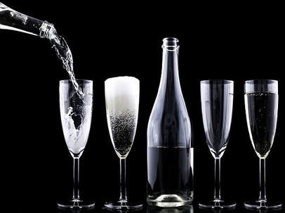 A short line up of flute glasses and bottle against a black background. To the left there is another bottle pouring fizzy drink into one of the flute glasses.