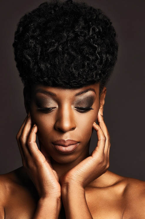 Black female model with a fancy updo. Her hands are y the side of her face and her eyes are looking down.