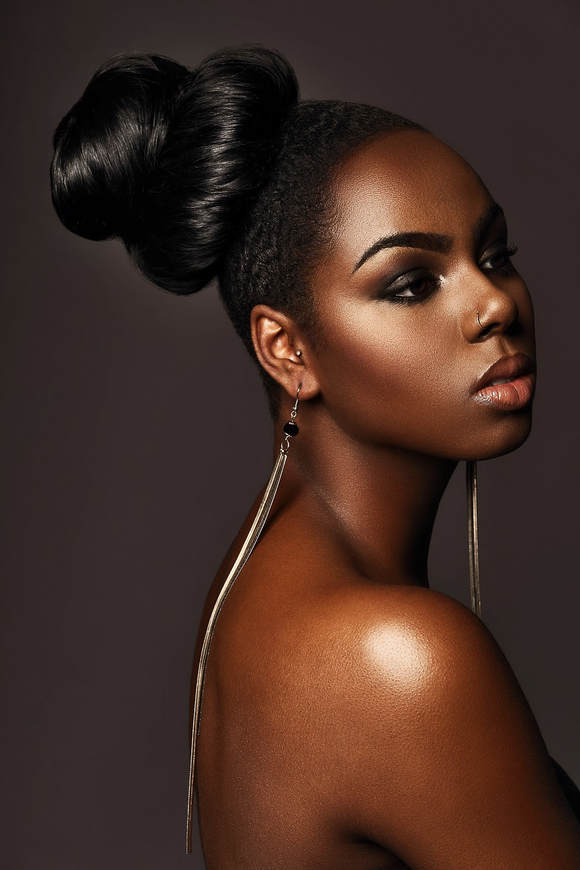 Black female model, modelling her hair. She is wearing a smooth looking updo and is wearing very long dangly earrings.