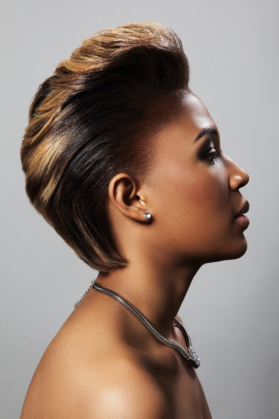 Black female, modelling her hair that has been cut short and is slicked back at the sides with a quif at the front. The hair has been been dyed a golden brown colour.